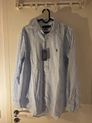  Ralph Lauren Skjorta - Klassisk långärmad skjorta från Polo Ralph Lauren i vitt och ljusblått randigt mönster. Skjortan har knappar framtill, krage och den ikoniska broderade loggan på bröstet. Tillverkad i bomull för en skön och luftig känsla. SMALL MEN MEDIUM I STORLEK. 
