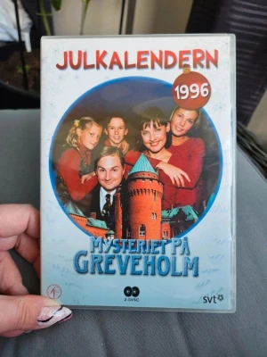 Julkalendern Mysteriet på Greveholm DVD - DVD-box med Julkalendern Mysteriet på Greveholm från 1996. Omslaget är färgglatt med bilder på karaktärerna och slottet. Innehåller 24 avsnitt på två skivor. Perfekt för dig som gillar nostalgi och klassiska svenska julkalendrar.