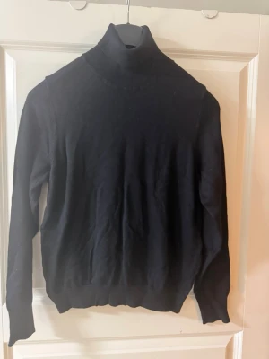 Svart polotröja stickad nyskick - Klassisk svart stickad polotröja med lång ärm och ribbade muddar. Tröjan har en hög polokrage och är tillverkad i ett mjukt, tunt material som passar perfekt till lager på lager. Enkel och stilren design som funkar till det mesta. I nyskick från ett djur och rökfri hem 