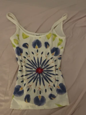 Blommig ribbad linnetopp Desigual - Säljer en vit ribbad linnetopp från Desigual med färgglatt blommigt mönster i blått, rött och gult. Toppen har smala axelband och är perfekt för dig som gillar unika och färgstarka plagg.