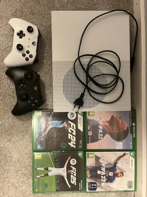 Xbox One S med två handkontroller - Säljer en vit Xbox One S med två trådlösa handkontroller, en vit och en svart. Konsolen är kompakt och stilren med ventilerad ovansida. Medföljer strömkabel och fyra spel: EA FC24, FIFA 22, FIFA 23 och FIFA 25. Perfekt för dig som gillar fotbollsspel! Kontrollen spakars gummiskydd är lite sönder på båda vänster spakar.