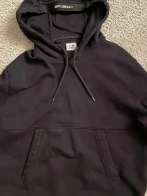 Svart hoodie från CP Company - Säljer en svart hoodie från CP Company med klassisk huva och dragsko. Hoodien har en stor magficka och en detaljficka med dragkedja på ena ärmen. Tillverkad i mjukt bomullsmaterial och har ribbade muddar vid ärmslut och nederkant.