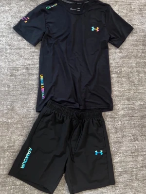 Svart träningsset från Under Armour - Säljer ett svart träningsset från Under Armour med t-shirt och shorts. Setet har färgglada och diskreta loggor på bröstet, ärmen och shortsen. Materialet är lätt och andas, perfekt för träning eller sportaktiviteter. Elastisk midja på shortsen för skön passform.  Säljer den för den inte passade mig köpte för 5 dagar sedan fick den för 2 dagar sedan säljer också för man inte kunde lämna tillbaka varan pris kan diskuteras 