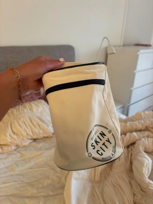 Beige necessär från Skincity - Säljer en beige necessär från Skincity med svart dragkedja och tryckt logga framtill. Väskan har cylinderform och ett praktiskt handtag upptill. Perfekt för att förvara smink eller hudvårdsprodukter. Materialet är troligtvis vattenavvisande syntet.