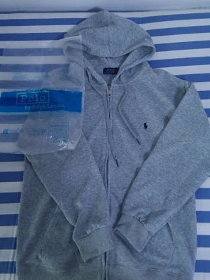 Grå hoodie från Polo Ralph Lauren - Snygg grå hoodie från Polo Ralph Lauren med dragkedja framtill och klassisk huva. Har det ikoniska broderade logotypen på bröstet och ribbade muddar vid ärmar och nederkant. Perfekt för en chill och stilren look.