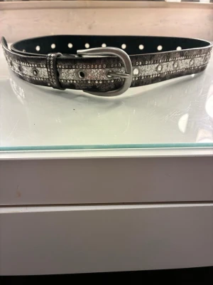 Svart & silvrigt Y2K strassbälte / skärp - Superfint svart skärp med silvriga detaljer, öljetter och glittriga strass-stenar! Ger den perfekta Y2K / grunge-looken till ett par snygga low rise jeans eller en kjol.                                              Skick: Oanvänt, men saknar prislapp!             Längd: 93 cm, bredd: 3 cm.