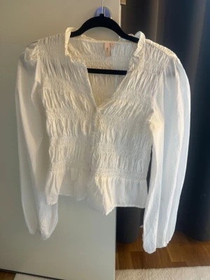 Vit smockad blus från Nelly - Säljer en vit blus från Nelly med smockade detaljer över bröstet och midjan. Blusen har lång ärm, lätt v-ringad hals och volangkant vid kragen. Tillverkad i ett tunt, luftigt tyg!