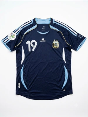 Argentina Adidas matchtröja blå - Snygg mörkblå Argentina matchtröja från Adidas med nummer 19, ljusblå och vita detaljer samt AFA-emblem på bröstet. Tröjan har korta ärmar, rund hals och klassiska Adidas-ränder på axlarna. Perfekt för fotbollsfans!