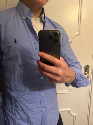 Ljusblå skjorta från Ralph Lauren  - Snygg ljusblå skjorta med klassisk knappkrage och broderad polohäst på bröstet. Skjortan har långa ärmar och knäppning framtill. Perfekt för en stilren look och passar till många olika outfits.