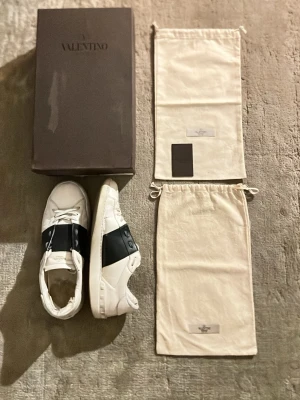 Vita sneakers med mörkblått band Valentino open - Snygga vita sneakers från Valentino med ett brett mörkblått band över mitten och diskret logga på tungan. Skorna har klassisk låg profil och vita snören. Kommer med originalkartong och två tygpåsar. Perfekta för dig som gillar stilrena och exklusiva sneakers.
