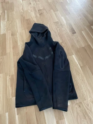 Svart hoodie med dragkedja och huva - Säljer en svart hoodie med hel dragkedja framtill och huva. Hoodien har en diskret design med reflexdetaljer över bröstet och en ficka med dragkedja på ena ärmen. Min kille vill inte ha kvar den nå mer i fint skick