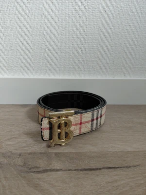 Burberry bälte  - Snyggt bälte i beige med klassiskt rutigt mönster och ett stort guldigt B-spänne framtill. Bältet har svarta och röda detaljer i mönstret och insidan är svart. Perfekt accessoar för att lyfta din outfit.