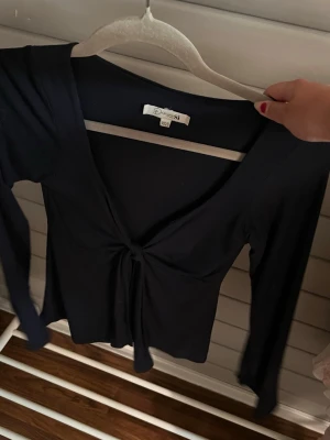Svart långärmad topp med knyt - Säljer en svart långärmad topp med snygg knytning framtill och djup v-ringning. Toppen har en slim passform och är perfekt för dig som gillar stilrena plagg med en twist. Passar till många olika looks och är superenkel att matcha.