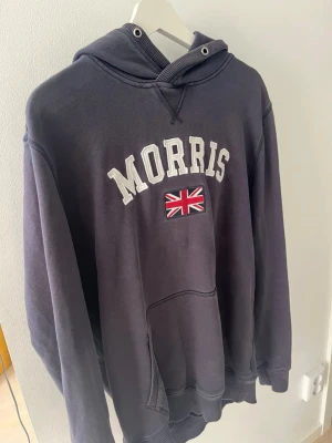 Mörkblå hoodie från Morris - Snygg mörkblå hoodie från Morris med stor vit text på bröstet och en broderad brittisk flagga under. Klassisk känguruficka framtill och huva med dragsko. Perfekt för en avslappnad och stilren look. Den är ganska urtvättad och saknar snören 