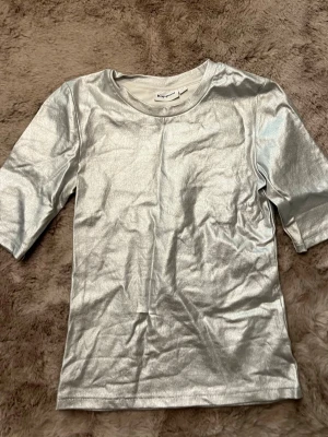 Metallic silver t-shirt från Kappahl - Snygg metallic silver t-shirt från Kappahl med rund halsringning och korta ärmar. Materialet har en glansig finish som ger en futuristisk vibe och sticker ut i mängden. Perfekt för dig som vill ha något unikt i garderoben.