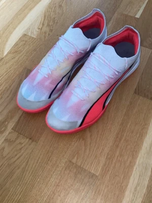 Vita Puma Ultra fotbollsskor - Säljer ett par snygga Puma Ultra fotbollsskor i vitt med rosa och svarta detaljer. Skorna har en lätt och ventilerande ovandel i syntetmaterial, snörning och en låg profil. Perfekta för dig som vill ha snabbhet och stil på planen.