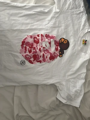Vit BAPE t-shirt med rosa camomönster - Säljer en vit t-shirt från BAPE med det klassiska apansiktet i rosa, rött och ljusrosa camomönster på bröstet. T-shirten har korta ärmar och en avslappnad passform. Perfekt för dig som gillar streetwear och vill sticka ut lite extra.