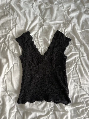 Svart spetsblus med v-ringning - Säljer en svart blus i spets med djup v-ringning och vågiga kanter. Toppen är ärmlös och har en figurnära passform som ger en feminin look. Perfekt att styla med jeans eller kjol för en trendig outfit.
