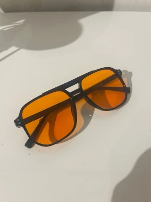 Svarta solglasögon med orange glas - Snygga solglasögon med svart båge och rektangulär form. Glasen är i en cool orange ton som verkligen sticker ut. Perfekta för dig som vill ha en unik och trendig accessoar till sommaren.