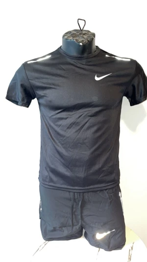 Svart träningsset från Nike - Snyggt svart träningsset från Nike med t-shirt och shorts. Båda plaggen har den klassiska vita Nike-loggan och är gjorda i ett lätt, snabbtorkande material som passar perfekt för träning eller löpning.