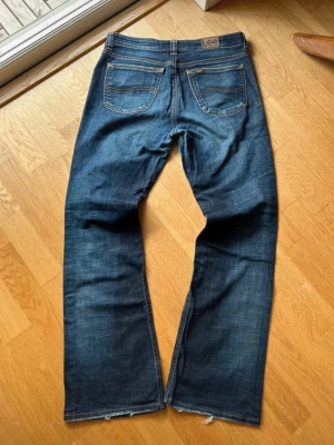Bootcut Jeans Lee - Säljer ett par klassiska blå jeans från Lee med rak passform och fem fickor. Jeansen har en snygg tvättad look, orange sömmar och råa kanter vid bensluten. Perfekta för en avslappnad stil och funkar till det mesta.
