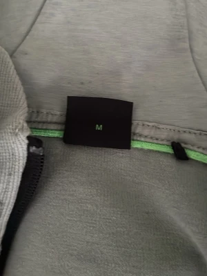 Ljusgrå hoodie med dragkedja - Säljer en ljusgrå hoodie med dragkedja och huva. Hoodien har svarta dragsnören och detaljer, samt fickor med dragkedja. Perfekt för chill dagar eller när du vill ha en enkel och clean look. Materialet känns mjukt och bekvämt.