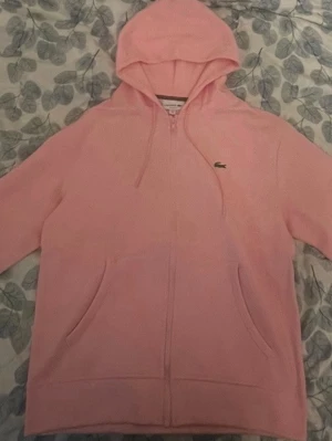 Lacoste hoodie - Säljer en ljusrosa hoodie från Lacoste i storlek M. Hoodien har dragkedja framtill, två fickor och justerbar huva med snören. Klassisk Lacoste-logga på bröstet. Tillverkad i mjukt bomullsmaterial för skön känsla.