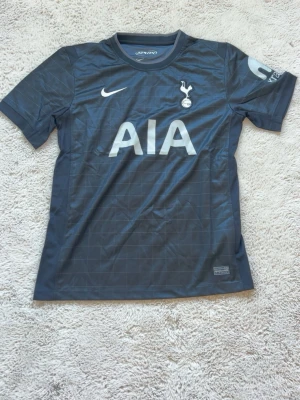 Tottenham Hotspur bortatröja Nike - Mörkgrå Tottenham Hotspur bortatröja från Nike med klubbmärke och AIA-logga på bröstet. Tröjan har korta ärmar, rund hals och diskret rutmönster i tyget. Tillverkad i lätt och ventilerande material, perfekt för fotbollsträning eller match. 🔴 pris kan diskuteras vid snabb affär🔴