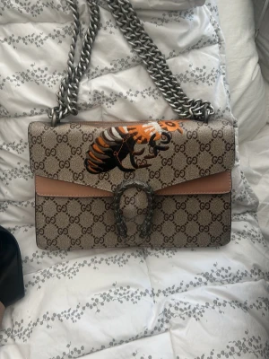 Gucci axelväska med bi-motiv - Säljer en snygg Gucci axelväska i beige med klassiskt GG-mönster och ett coolt bi-motiv på locket. Väskan har en kraftig silverfärgad kedja och detaljer i ljusbrunt läder. Perfekt för dig som vill sticka ut med en unik designerbag.