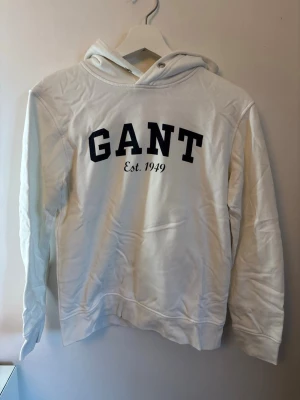Vit hoodie från GANT  - Snygg vit hoodie från GANT med svart text 'GANT Est. 1949' på bröstet. Klassisk modell med huva och dragsko, ribbade muddar vid ärmslut och nederkant. Perfekt för en avslappnad och stilren look.
