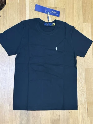Svart t-shirt från Polo Ralph Lauren - Klassisk svart t-shirt från Polo Ralph Lauren med rund halsringning och korta ärmar. Diskret vit logobrodyr på bröstet. Tillverkad i mjuk bomull för en skön och stilren look.
