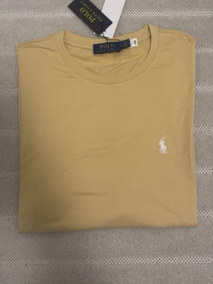 Beige t-shirt från Polo Ralph Lauren - Stilren beige t-shirt från Polo Ralph Lauren med klassisk rund halsringning och ikonisk vit broderad logga på bröstet. Tillverkad i mjuk bomull som känns skön mot huden. Perfekt basplagg för en clean och avslappnad look.