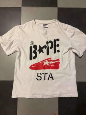 Vit Bape STA t-shirt med tryck - Säljer en vit t-shirt från Bape med stort svart och rött tryck på framsidan. Motivet visar BAPE-loggan och en röd sneaker med stjärna. Klassisk rund hals och korta ärmar. Perfekt för dig som gillar streetwear och vill sticka ut.