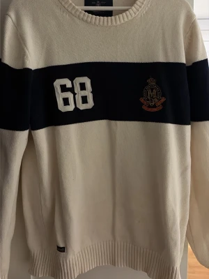 Vit stickad tröja med marinblått band - Säljer en vit stickad tröja med ett brett marinblått band över bröstet, siffran 68 i vitt och ett broderat emblem. Tröjan har rund halsringning och långa ärmar. Perfekt för dig som gillar sportig och klassisk stil.