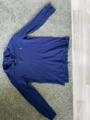 Mörkblå långärmad Ralph Lauren Polo piké - Mörkblå långärmad Ralph Lauren Polo piké i mjuk bomull med klassisk krage och knappslå framtill. Pikén har en liten broderad logga på bröstet (Ralph Lauren Polo loggan) och en enkel, stilren design som funkar till det mesta.