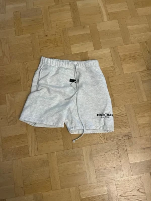 Gråa shorts från Essentials - Snygga ljusgrå shorts från Essentials med elastisk midja och snörning. Mjukt material och logga på ena benet. Perfekta för chill dagar eller när du vill ha en clean look.