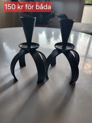Svarta metall ljusstakar, 2-pack - Två stilrena ljusstakar i svart metall med modern, industriell design. De har tre böjda ben och en konformad hållare för stearinljus. Perfekta som inredningsdetalj på bord eller hylla. 