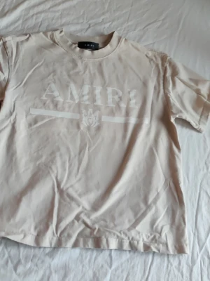 Beige t-shirt från AMIRI - Säljer en beige t-shirt från AMIRI med tryckt logga framtill. T-shirten har klassisk passform, rund halsringning och korta ärmar. Perfekt för dig som gillar stilrena och trendiga plagg.