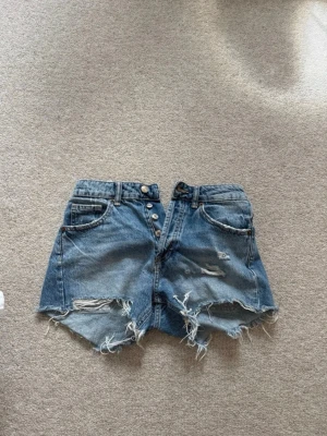 Blå slitna jeansshorts med knappar - Säljer ett par blå jeansshorts med hög midja och råa, fransiga kanter. Shortsen har flera slitningar framtill, knappgylf och klassiska fem fickor. Perfekta för sommaren och ger en avslappnad, trendig look.