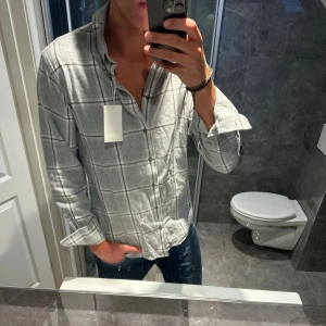 Grå rutig skjorta med knappar - Riktigt schysst ljusgrå flanellskjorta från Bruun & stengade. Skjortan har aldrig använts, prislapp sitter kvar. Köptes ny för 800kr. Jag på bilden är 188cm och bör strlk M. Hör av er vid eventuella frågor.