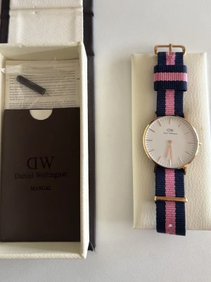 Daniel Wellington klocka randig blå/rosa - Snygg Daniel Wellington klocka med vit urtavla och guldfärgade detaljer. Armbandet är randigt i marinblått och rosa, gjort i textil. Klassisk och stilren design som passar till många olika looks. Är i behov av batteribyte. Plastskyddet på baksidan är kvar på klockan Levereras med originalask och manual.