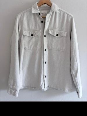 Ljusgrå overshirt från Jack & Jones - Stilren ljusgrå overshirt från Jack & Jones med två bröstfickor med lock och svarta knappar. Overshirten har klassisk krage och är tillverkad i ett mjukt, strukturerat tyg som ger en avslappnad men snygg look.