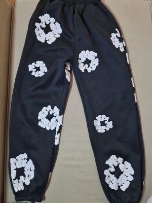 Svarta mjukisbyxor med vita blommor - Säljer ett par svarta mjukisbyxor med vita blommönster. Byxorna har resår i midjan och vid bensluten för en skön och avslappnad passform. Perfekta för chill dagar eller när du vill ha något bekvämt på dig.