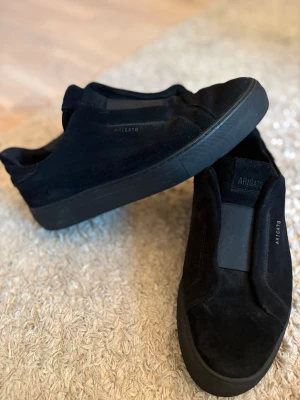 Svarta sneakers från Axel Arigato - Stilrena svarta sneakers från Axel Arigato i mocka med diskret logga på sidan och plösen. Skorna har en låg silhuett och svart sula i gummi. Perfekta för dig som gillar minimalistisk och clean design.