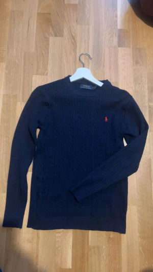 Mörkblå kabelstickad tröja Polo Ralph Lauren - Mörkblå kabelstickad tröja från Polo Ralph Lauren med rund halsringning och röd broderad logga på bröstet. Tröjan har långa ärmar och ribbade muddar vid ärmslut och nederkant. Perfekt för dig som gillar klassisk och stilren design.