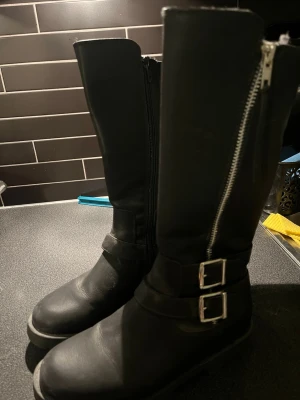 Svarta höga stövlar med spännen - Snygga svarta stövlar i skinnimitation med högt skaft, fodrade, grov sula och två dekorativa spännen i silver. Dragkedja på utsidan av skaftet för en cool look. Perfekta för dig som gillar en tuffare stil. Helt oanvända💗