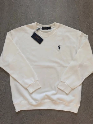 Vit sweatshirt från Polo Ralph Lauren - Klassisk vit sweatshirt från Polo Ralph Lauren med rund halsringning och diskret logobrodyr på bröstet. Tröjan har långa ärmar och ribbade muddar vid ärmslut och nederkant. Perfekt basplagg för en clean och stilren look.
