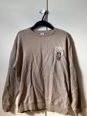 Beige sweatshirt med bi-tryck - Cool beige sweatshirt med tryck av en färgglad bi på bröstet och ryggen. Textdetaljer på ryggen i vitt och gult. Tröjan har rund halsringning och långa ärmar. Perfekt för dig som gillar unika prints och avslappnad stil. Storlek 13-14år/164cm