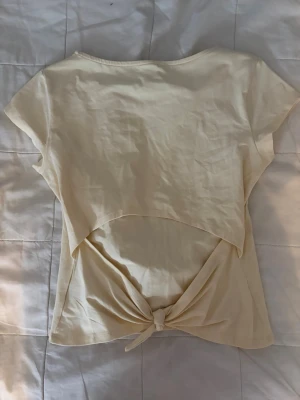 Gul topp med knytning bak - Snygg beige topp i mjukt bomullstyg med korta ärmar och rund halsringning. Baksidan har en öppen detalj med knytning nedtill som ger en trendig look. Perfekt att matcha med jeans eller kjol för en avslappnad stil.