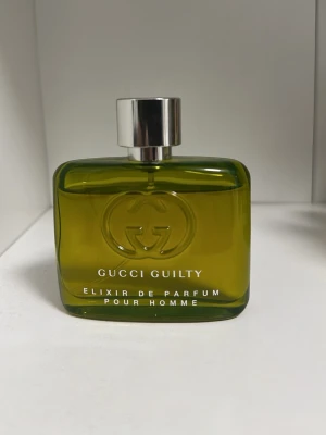 Gucci Guilty Elixir Pour Homme parfym - Gucci Guilty Elixir de Parfum Pour Homme i en stilren, rektangulär flaska av grönt glas med silverfärgad kork. Flaskan har Gucci-loggan ingraverad på framsidan och text i vitt. En exklusiv herrdoft som sticker ut i badrumshyllan.